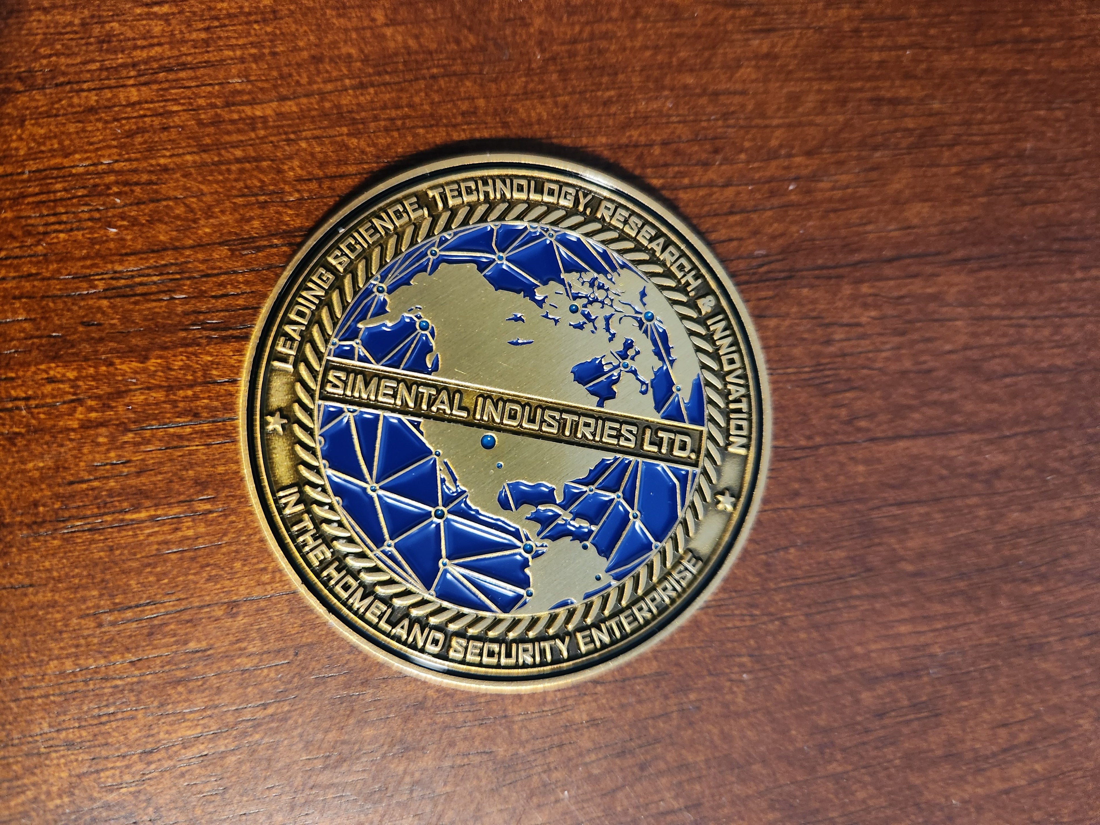 Close-up of the Limited Edition Disaster Gaming Challenge Coin front side showing a blue world map design and 'Simental Industries Ltd.' banner on a wooden surface